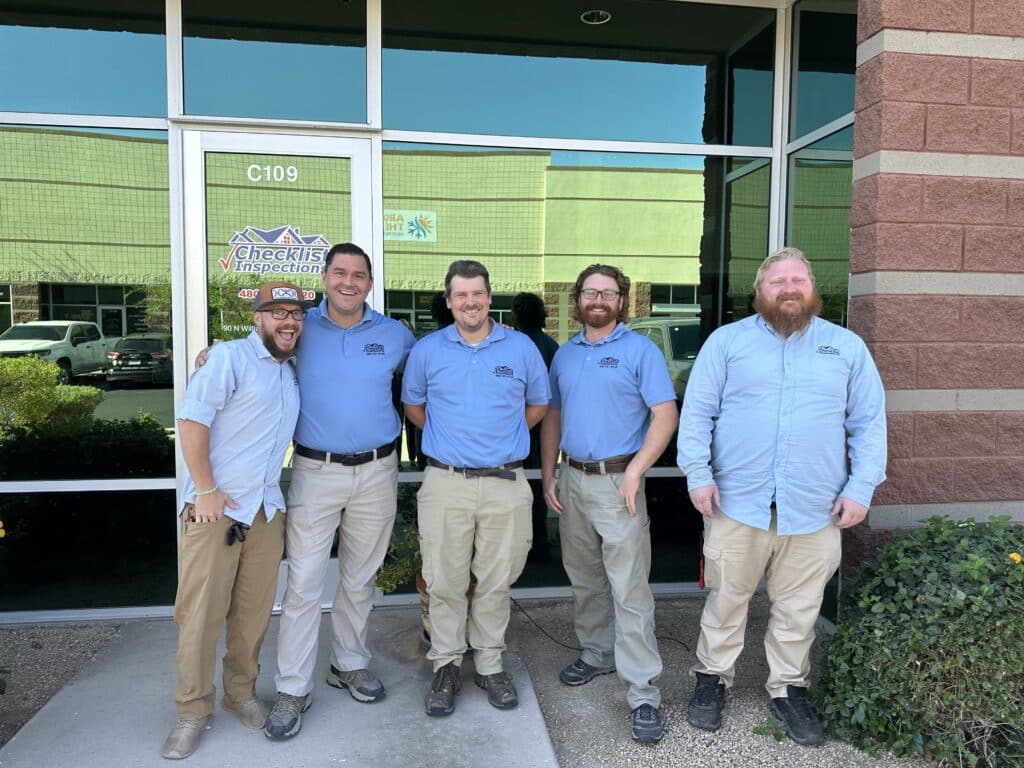 Five men in matching light blue shirts and khaki pants smile together outside a glass building entrance with a "Checklist Inspections" logo.