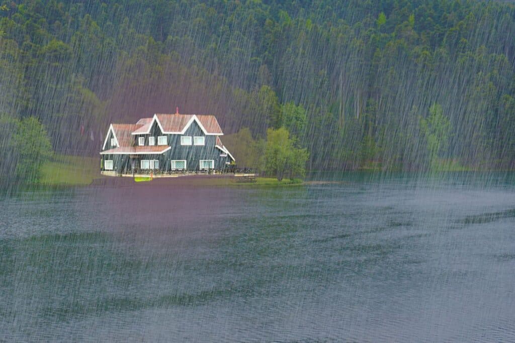 A multi-story lodge-style house on the edge of a large lake during a heavy rainstorm, backed by a dense forest.