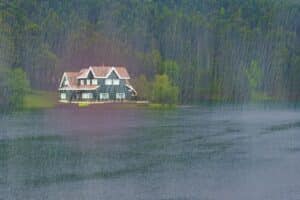 A multi-story lodge-style house on the edge of a large lake during a heavy rainstorm, backed by a dense forest.